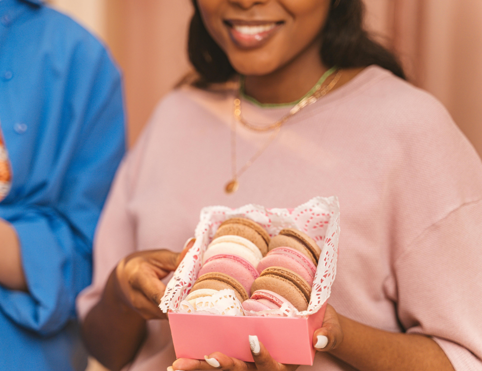 Smiling girl wearing pink, holding a pink box of pink, white and brown macarons.